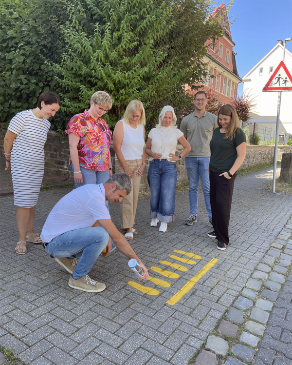 Bild vergr&ouml;&szlig;ern: Gelbe F��e werden f�r die Sicherheit der Kinder aufgemalt. Foto Stadt Lennestadt, v.l.n.r. Sandra Friedrichs von der Elternvertretung, Mobilit�tsmanager Philip Laister (hockend), Schulbereichsleiterin der Stadt Lennestadt Britta He�, Schulleiterin Karin Brieden, Polizeibeamtin Kerstin Brauer, Kevin Schulte vom Ordnungsamt der Stadt Lennestadt und Marina Artino von der Elternvertretung.