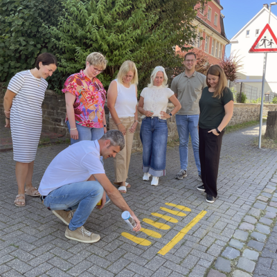 Gelbe F��e werden f�r die Sicherheit der Kinder aufgemalt. Foto Stadt Lennestadt, v.l.n.r. Sandra Friedrichs von der Elternvertretung, Mobilit�tsmanager Philip Laister (hockend), Schulbereichsleiterin der Stadt Lennestadt Britta He�, Schulleiterin Karin Brieden, Polizeibeamtin Kerstin Brauer, Kevin Schulte vom Ordnungsamt der Stadt Lennestadt und Marina Artino von der Elternvertretung. 
